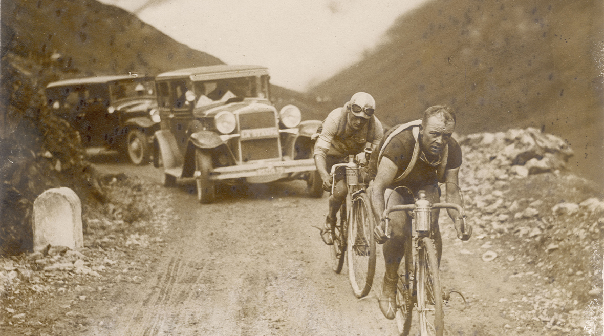 Two cyclists looking very tired riding up a mountain pass followed by two cars. An old black and white image.
